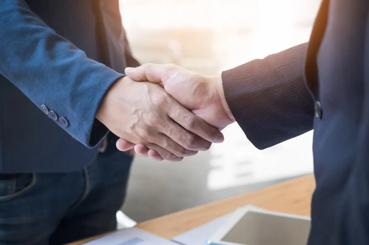Two people in suits shaking hands across a desk